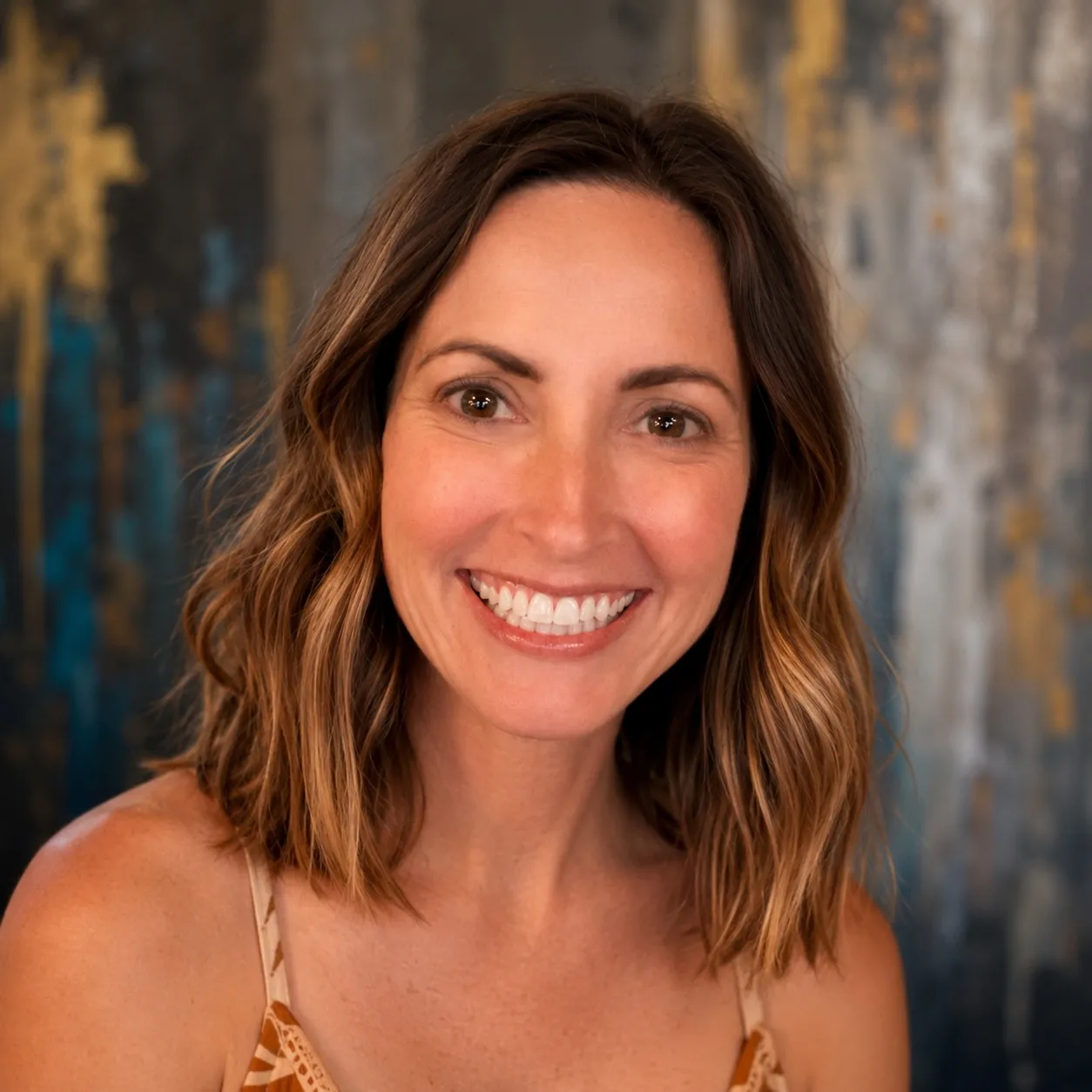Cathy Walls smiling at the camera, shoulder-length wavy brown hair with sun-lightened ends, in a rust-orange camisole. Behind her hangs one of her own paintings — a tall abstract in slate grey and deep teal, streaked and dripped like weathered metal, with a burst of gold leaf at the upper left.